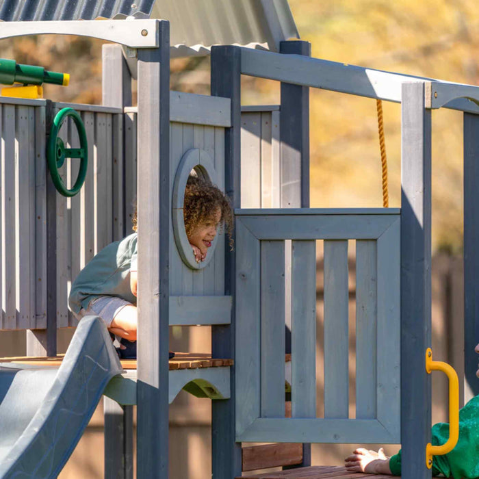 Jack and June Haven 2 Outdoor wooden playset with swing and slide, climbing ramp with rope and undercover landing. View of child putting her head out the window